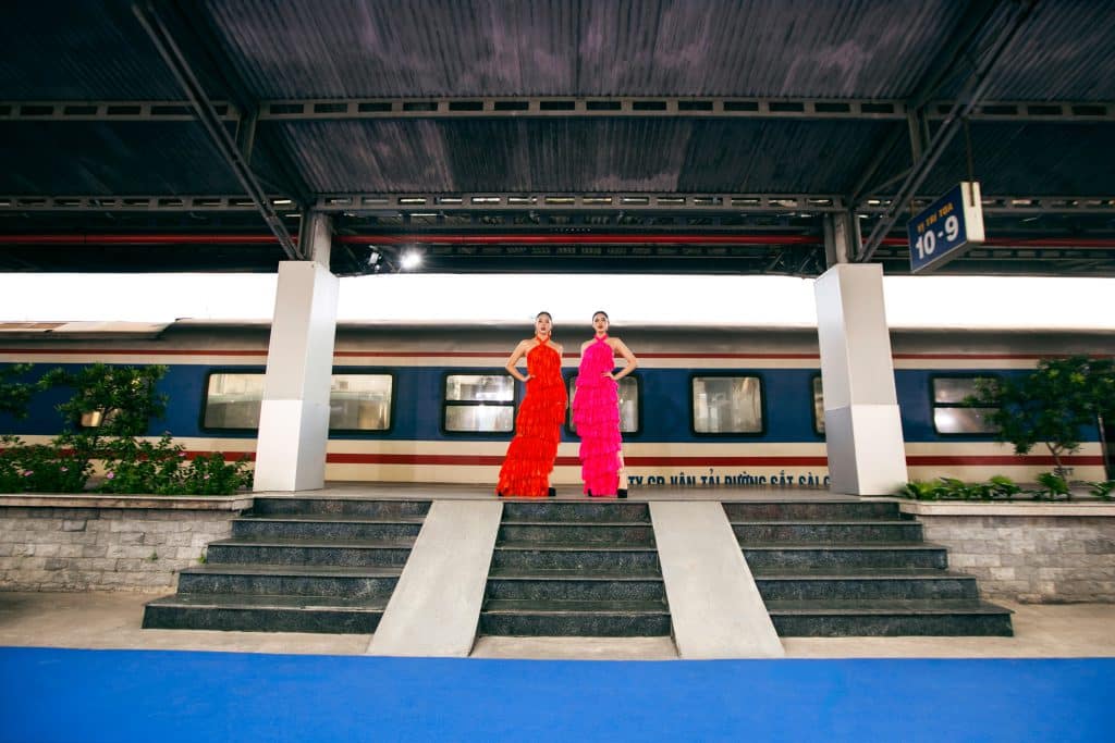 Hai người mẫu diện váy đỏ và hồng trên sân ga tàu hỏa