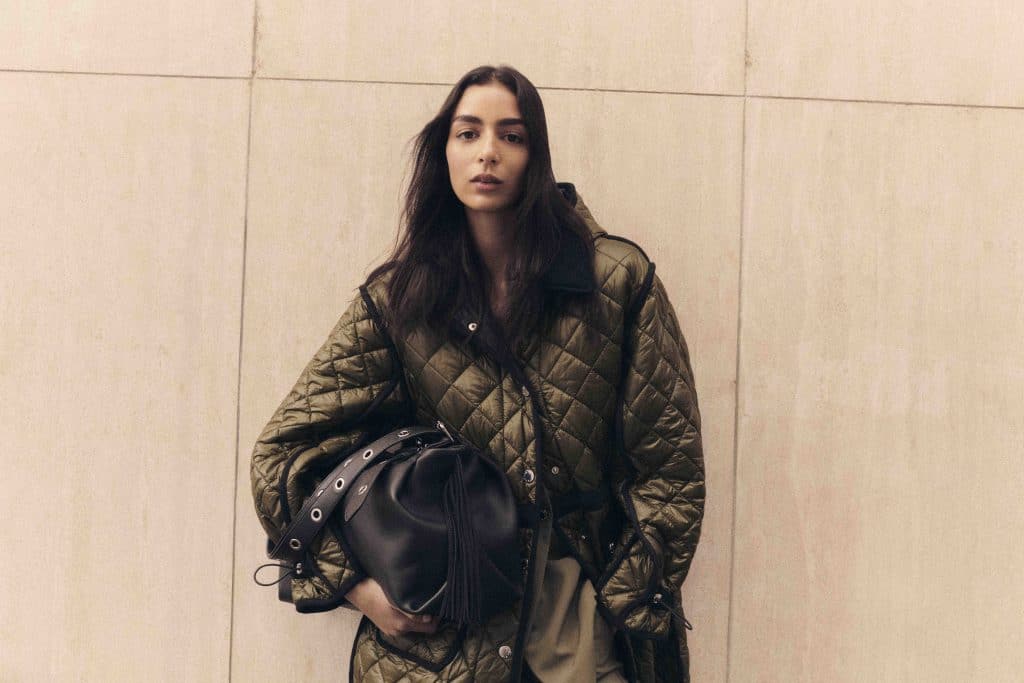 Người mẫu mặc áo khoác phồng và cầm túi xách da màu đen trước nền tường sáng màu