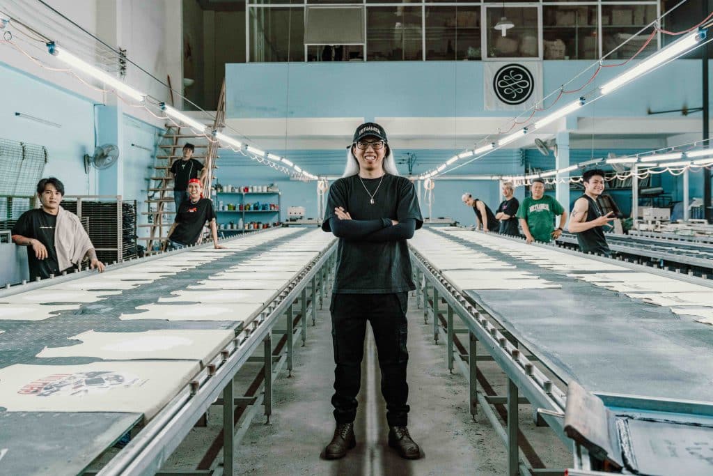 Người đàn ông trẻ đứng giữa xưởng in áo phông rộng lớn với nhiều nhân viên đang làm việc