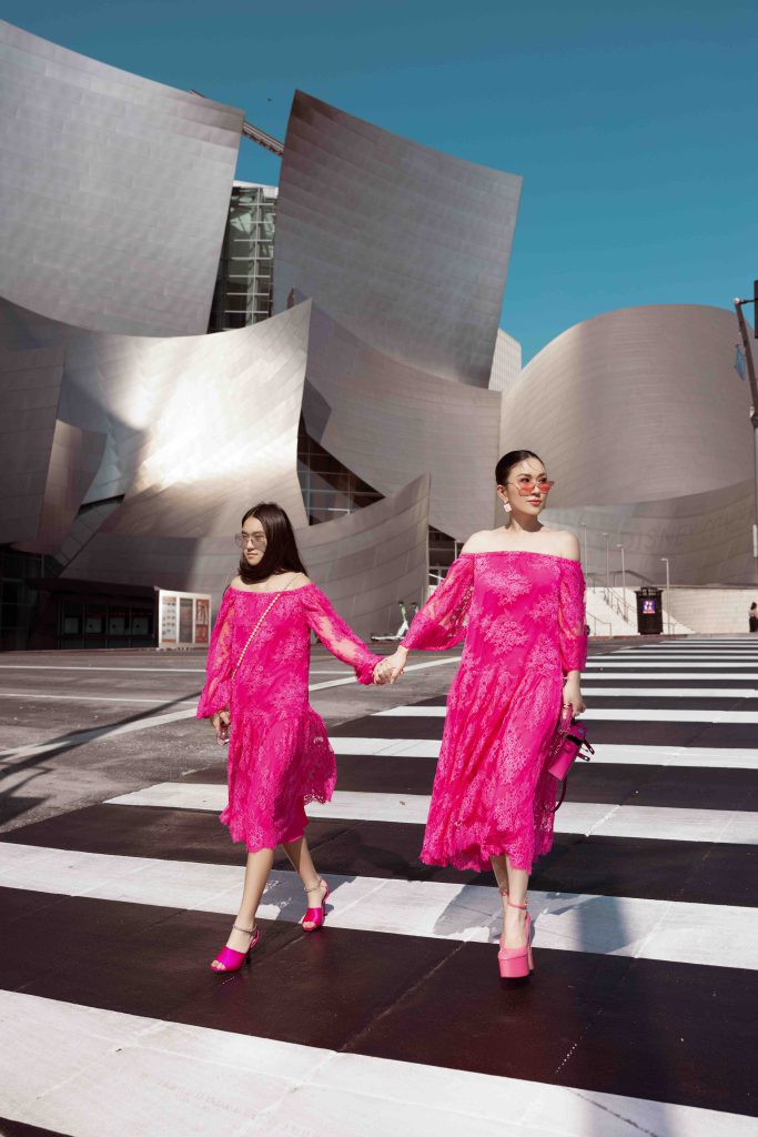 Mẹ và con gái diện đồ đôi màu hồng tươi tắn đi qua vạch kẻ đường trước một tòa nhà hiện đại