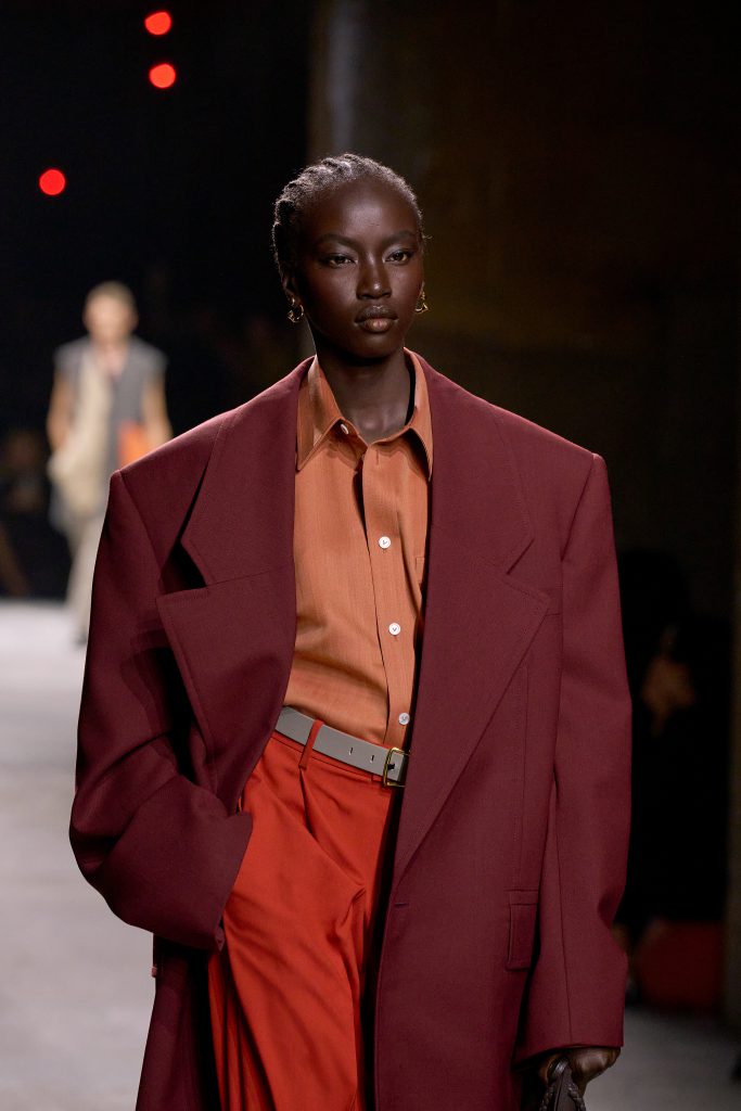 Người mẫu trình diễn bộ trang phục gồm áo sơ mi cam và áo blazer đỏ burgundy trên sàn diễn thời trang