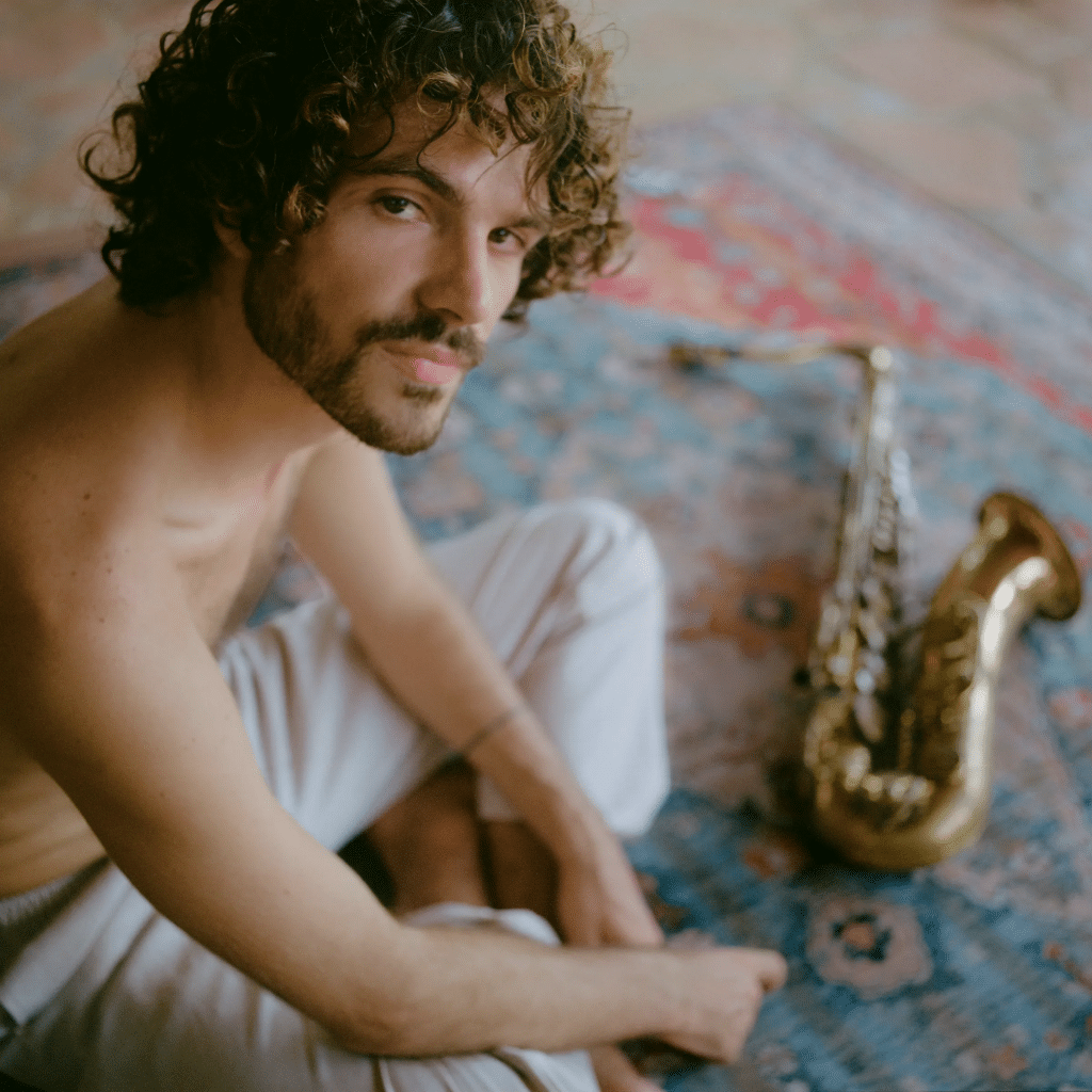 Người đàn ông trẻ với mái tóc xoăn ngồi cạnh chiếc kèn saxophone vàng trên thảm họa tiết