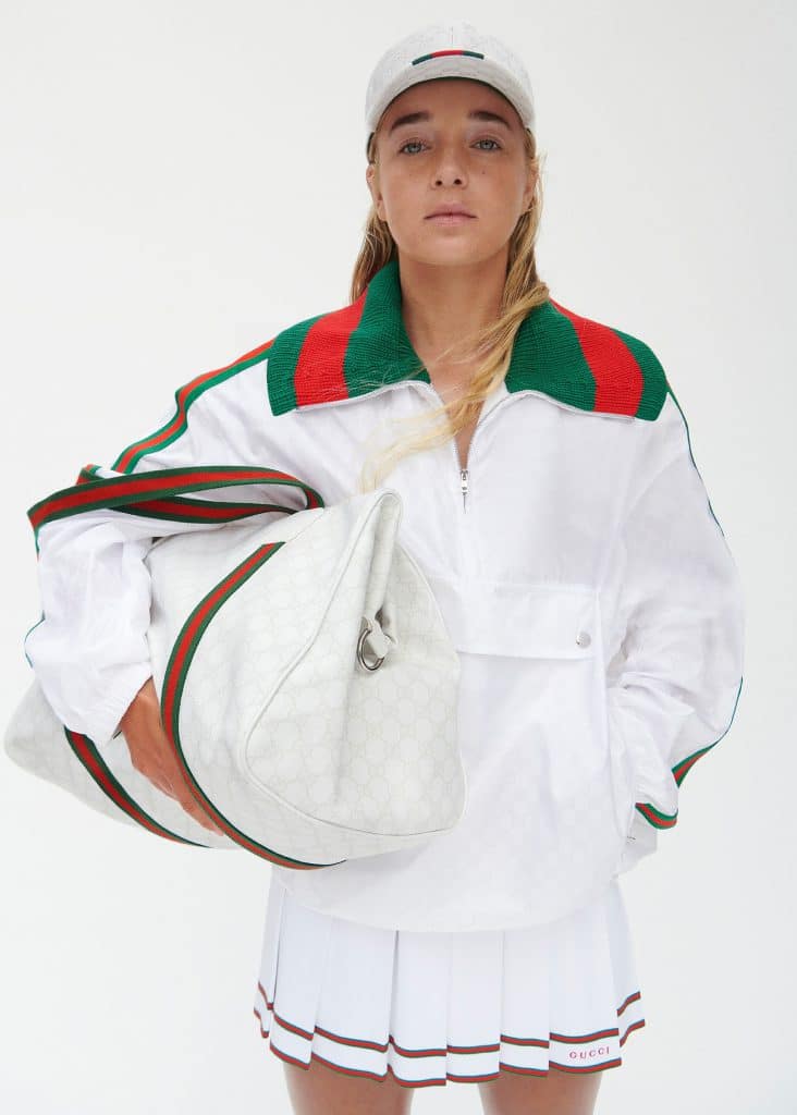 A model poses in white Gucci athletic wear with green and red stripes holding a large patterned bag