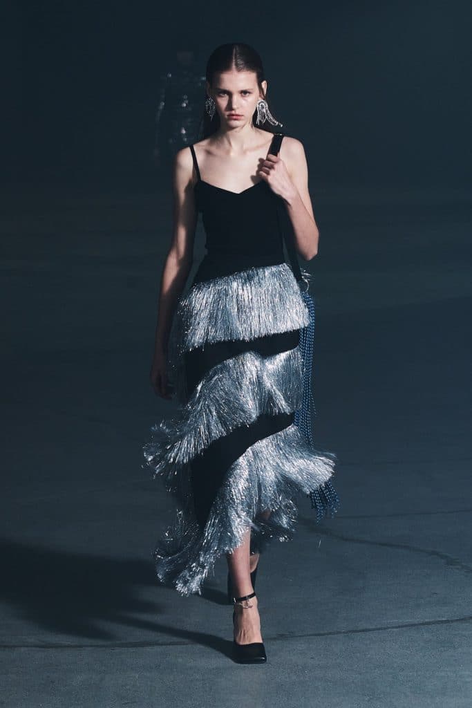 A model walks the runway wearing a black dress with a shimmering silver fringe skirt
