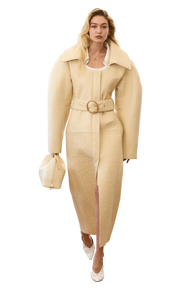 A model walks wearing a long beige coat with large sleeves and a textured skirt