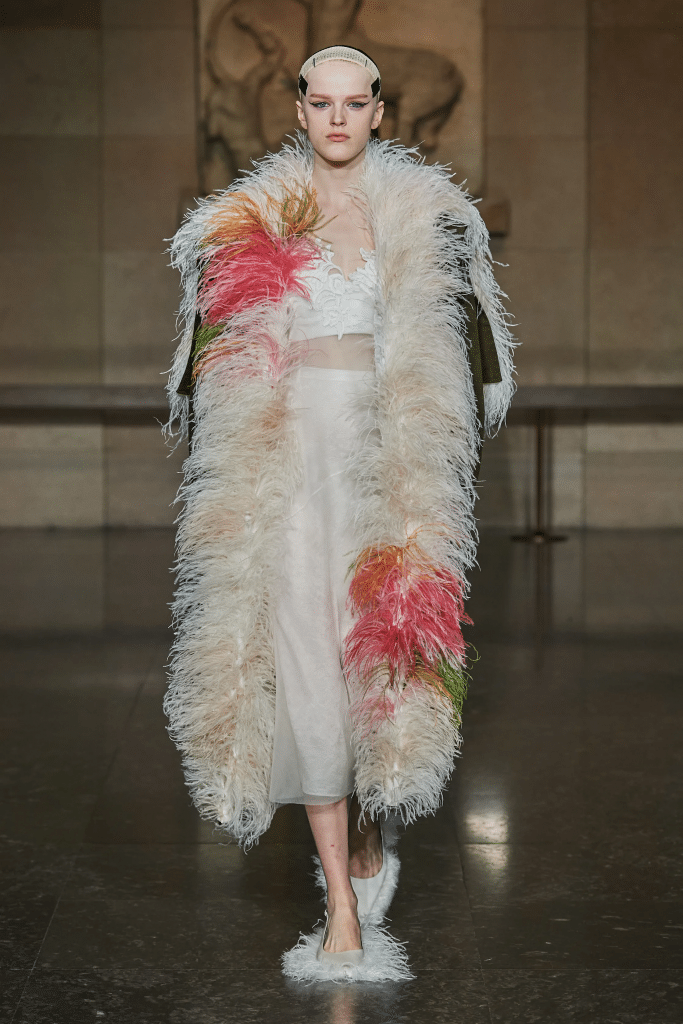 A model walks a runway wearing a long feathered coat and a light dress