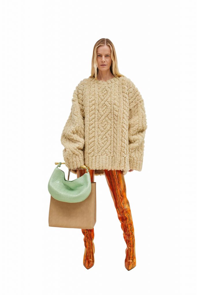 Model wears a chunky knit sweater orange pants and carries a green handbag