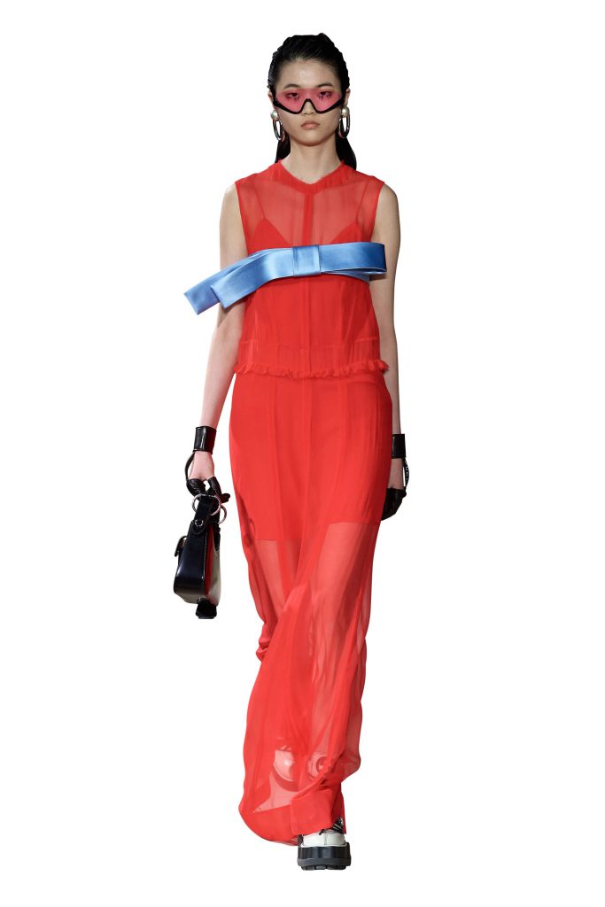A model walks wearing a red sheer dress blue sash and futuristic pink goggles