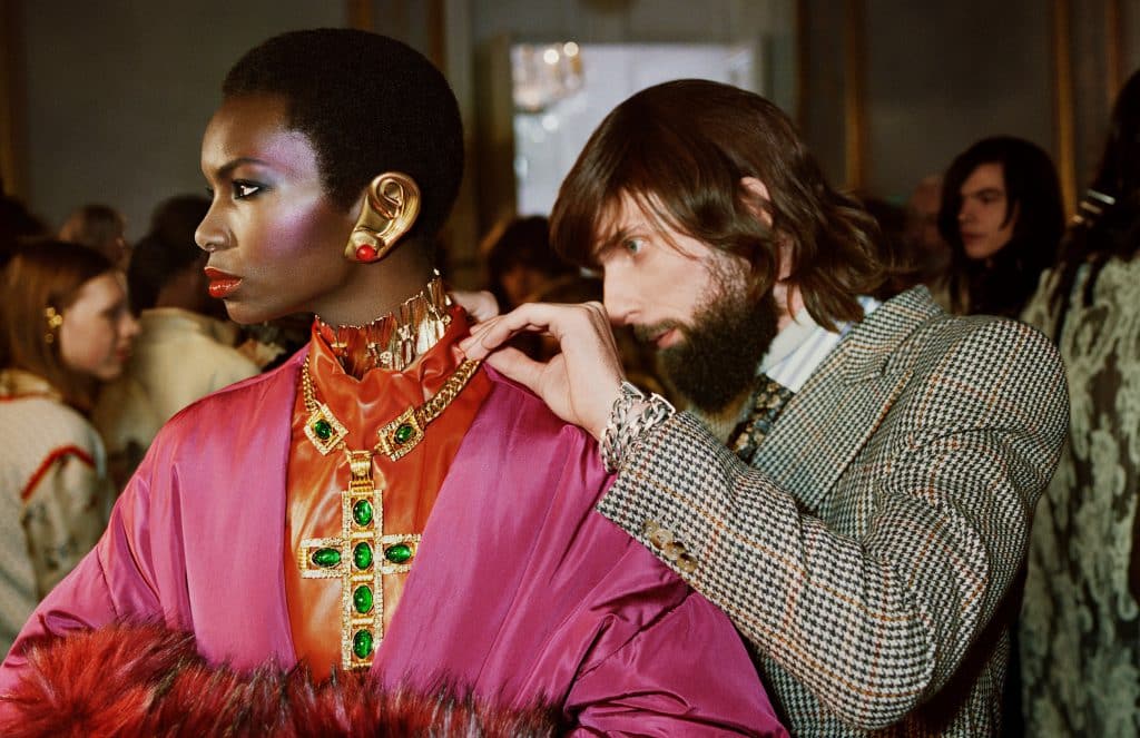A model wears bold jewelry while a stylist adjusts her hair backstage