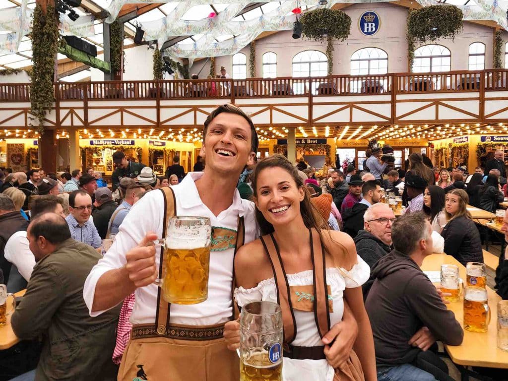 A smiling couple holds large beer steins at a crowded Oktoberfest celebration