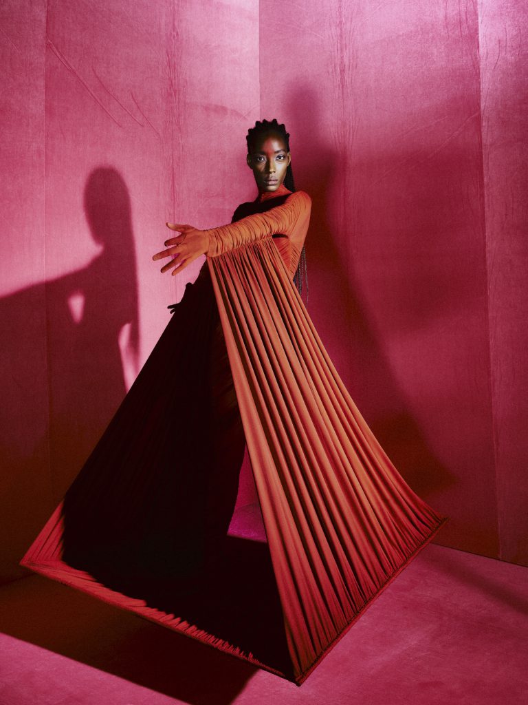 Người mẫu da màu mặc váy đỏ cam dáng dài tạo dáng trong không gian ánh sáng hồng
