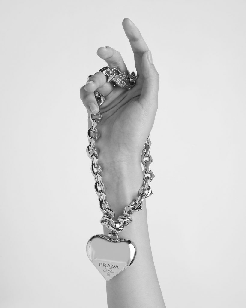 Một bàn tay cầm chuỗi vòng cổ và nhẫn kim loại sáng bóng thương hiệu Prada