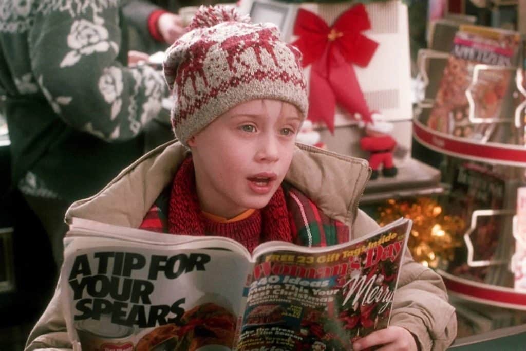 A young boy reads a newspaper wearing a winter hat and sweater near gifts