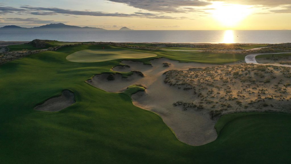 A lush green golf course meets sandy dunes near the ocean at sunset