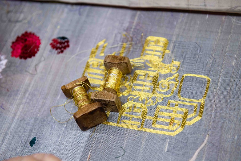 Gold thread and wooden spools rest on embroidered fabric near red flower details