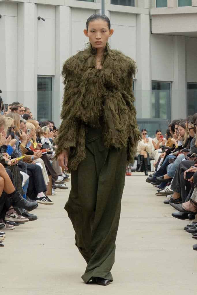 A model walks a runway wearing an olive green textured jacket and wide leg pants before an audience