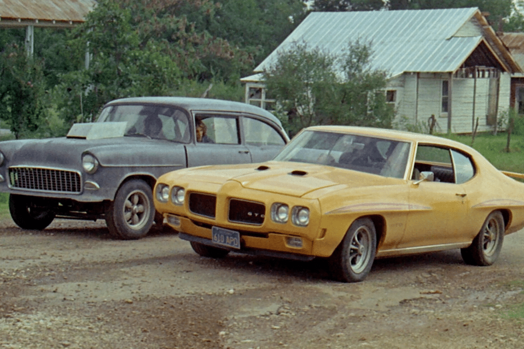 Hai chiếc xe ô tô cổ điển đỗ trước một ngôi nhà gỗ đơn sơ