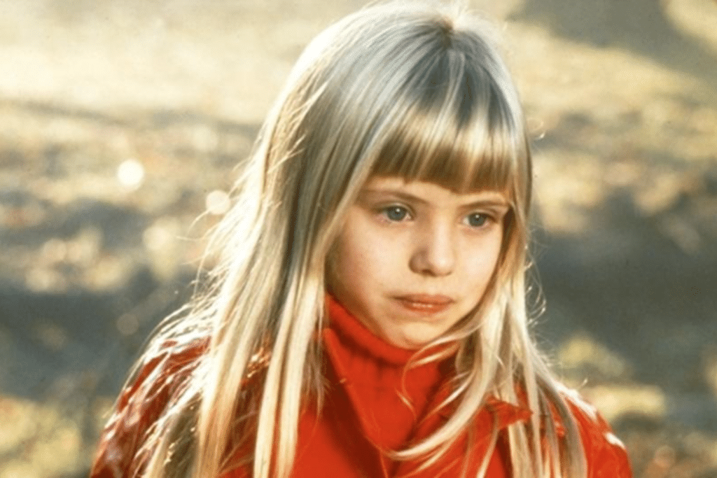 Cô bé với mái tóc vàng và áo cổ lọ đỏ nhìn thẳng vào ống kính