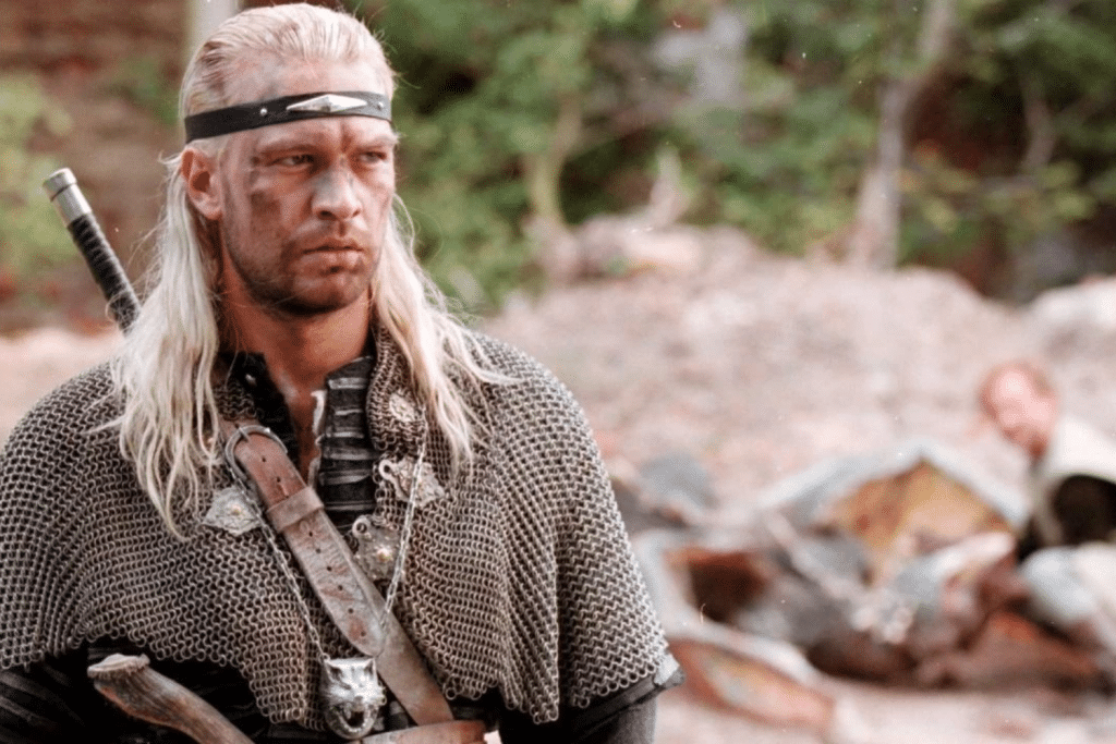 Một người đàn ông với tóc dài mặc áo giáp lưới và đeo băng đô đen đang nhìn xa xăm