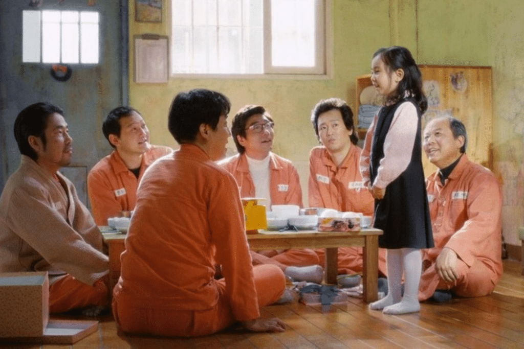 Một bé gái đứng trước nhóm tù nhân mặc áo cam đang ngồi ăn trong phòng giam