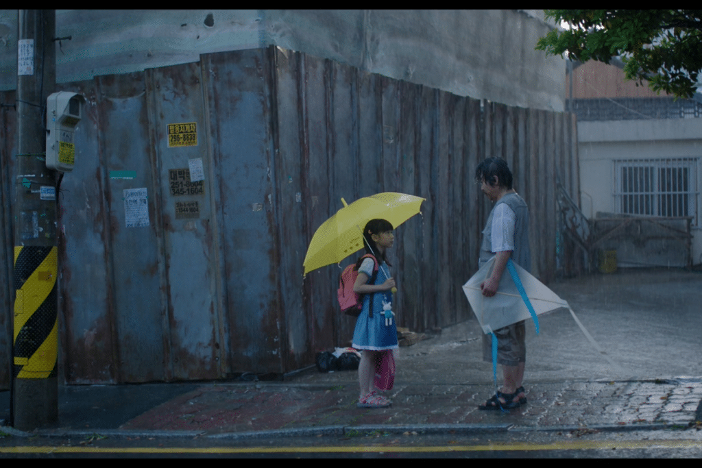 Bé gái cầm ô vàng đứng cạnh người đàn ông dưới mưa gần bức tường cũ