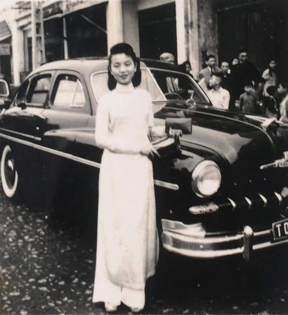 A bride poses in a white ao dai next to a classic black car with onlookers nearby