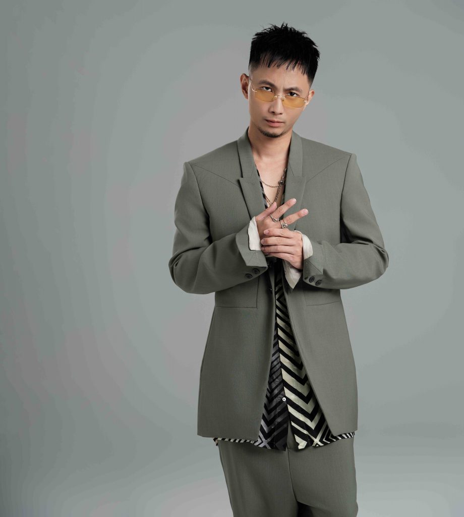 Một người đàn ông mặc vest và áo sơ mi kẻ sọc đang tạo dáng trước nền xám