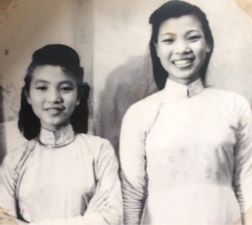 Two young women smile wearing traditional áo dài dresses from the 1940s