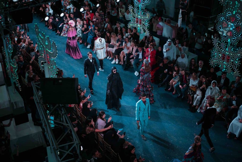 Models walk a blue runway before a seated audience at a fashion show