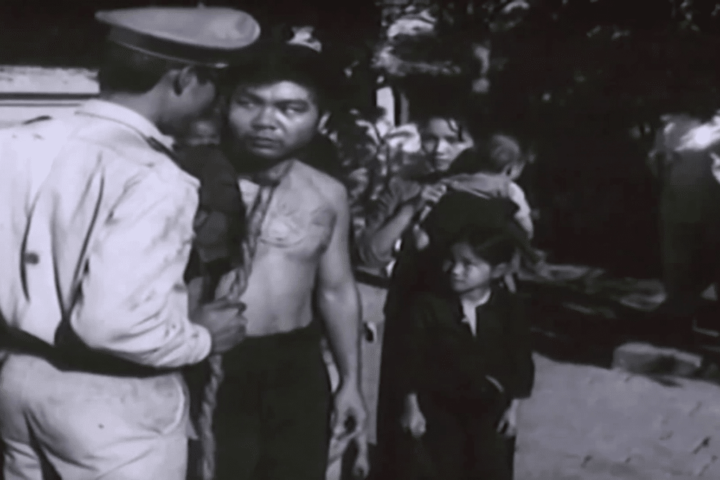 Cảnh phim đen trắng với nhân vật bị tra tấn và những người xung quanh