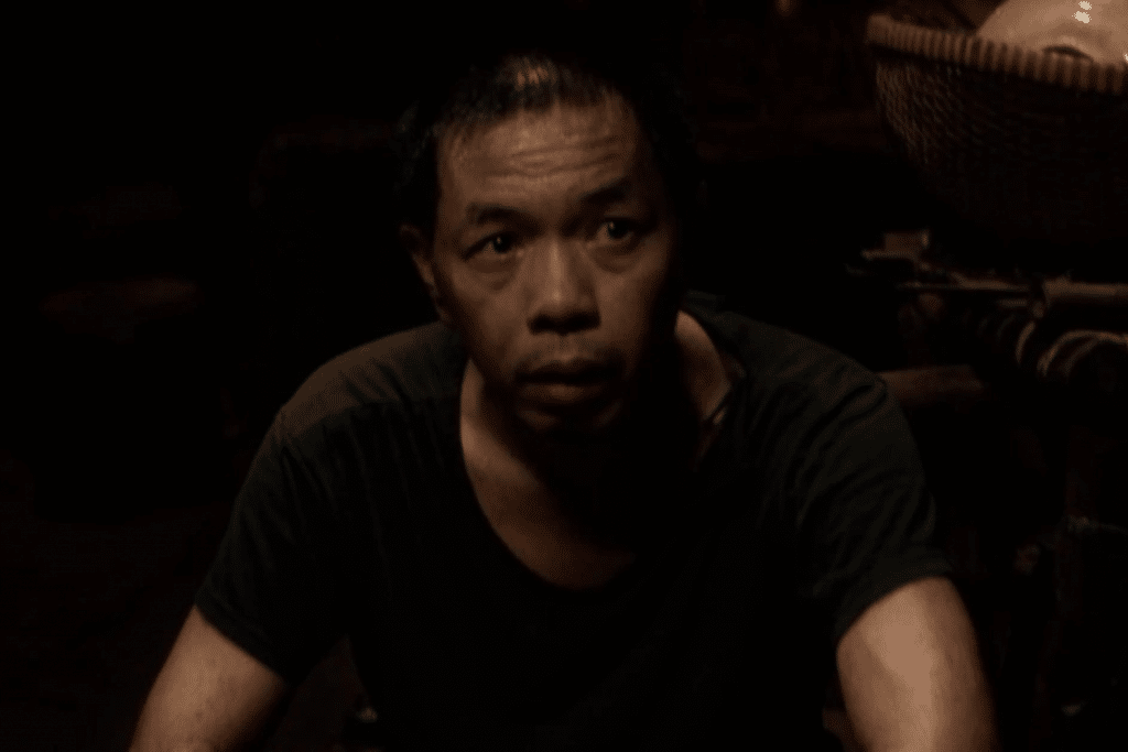 Một người đàn ông với biểu cảm căng thẳng nhìn thẳng vào ống kính trong khung cảnh tối tăm