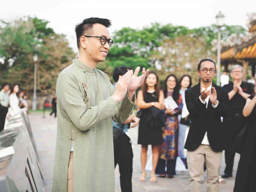 Người đàn ông mặc áo dài xanh lá đang vỗ tay cùng mọi người ở ngoài trời