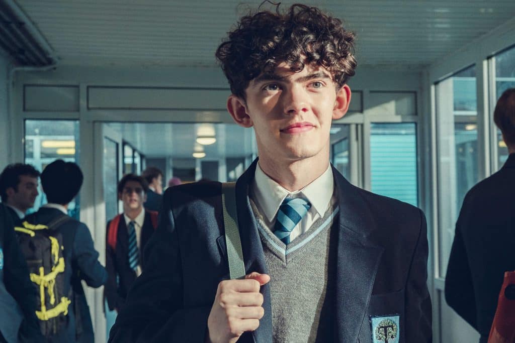 Nam sinh với mái tóc xoăn đang mỉm cười nhìn thẳng vào ống kính trong hành lang trường học