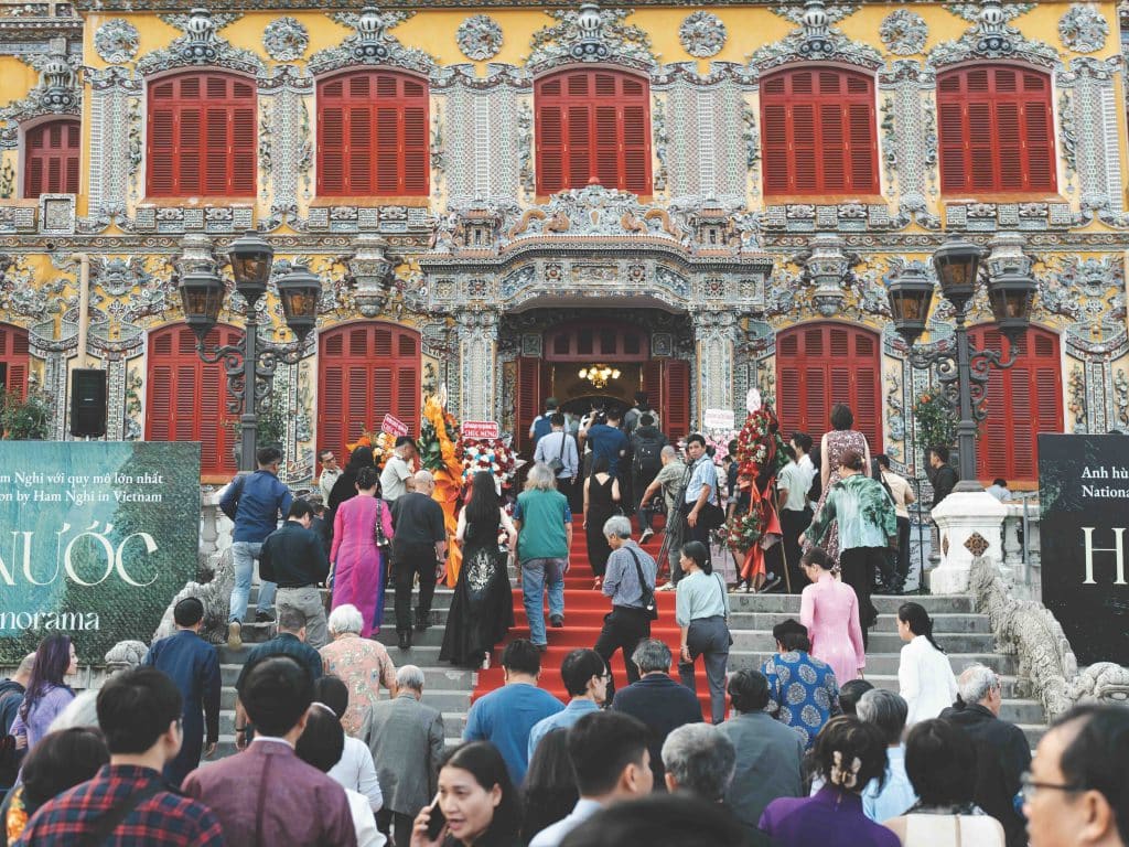 A large crowd gathers outside an ornate yellow building with red window shutters