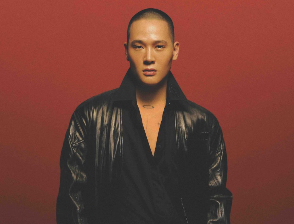 A man with a shaved head wears a black leather jacket against a red background