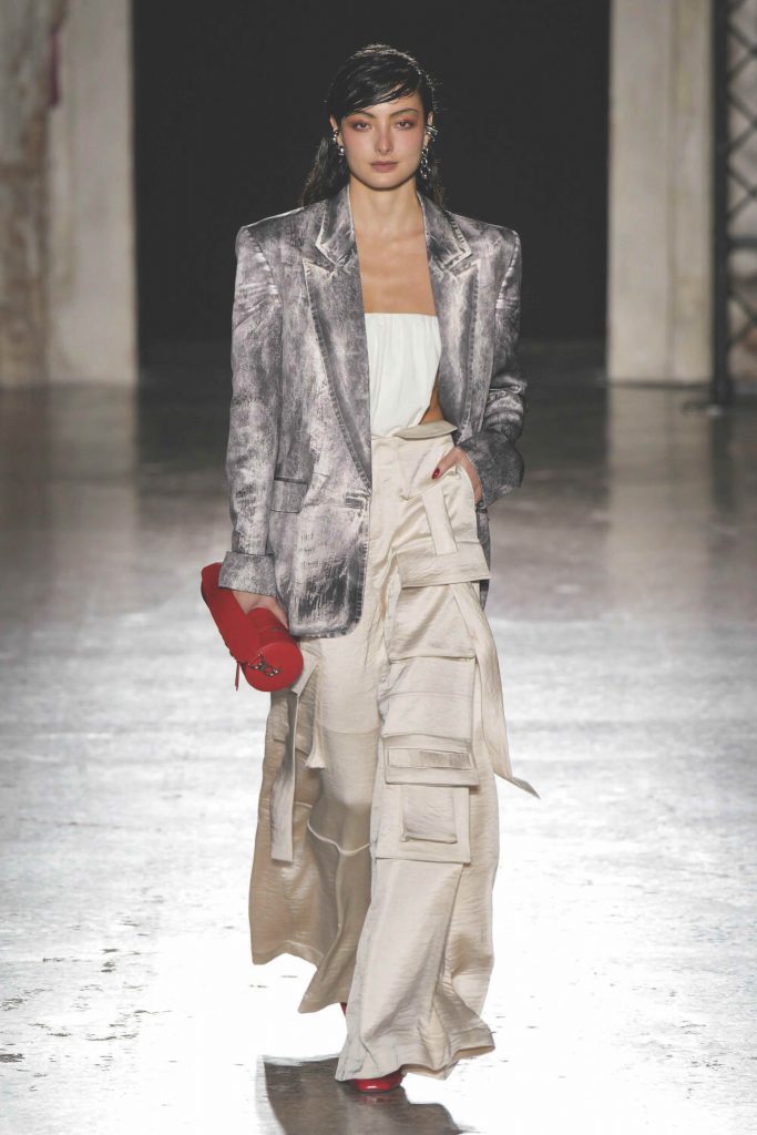 A model walks a runway wearing a blazer wide pants and carrying a red heart shaped bag