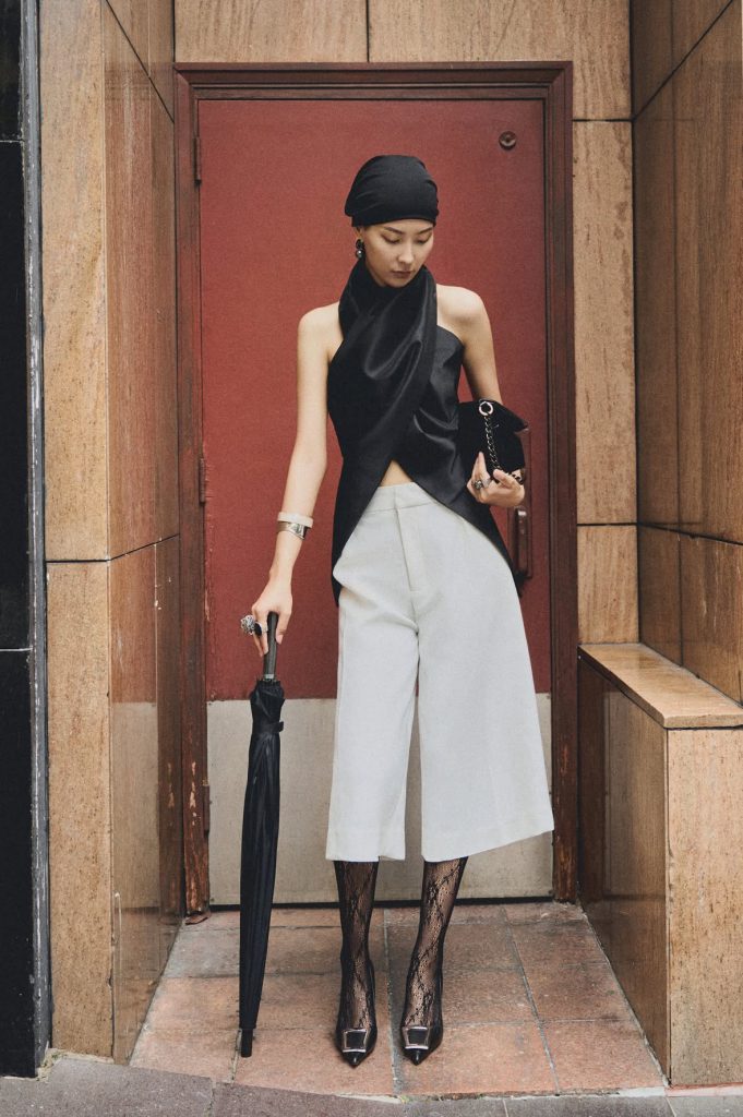 A woman in a black headwrap and stylish outfit holds a closed black umbrella