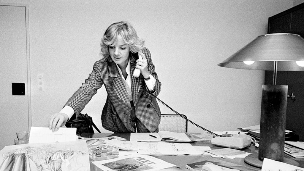A woman with blonde hair talks on the phone at a cluttered desk