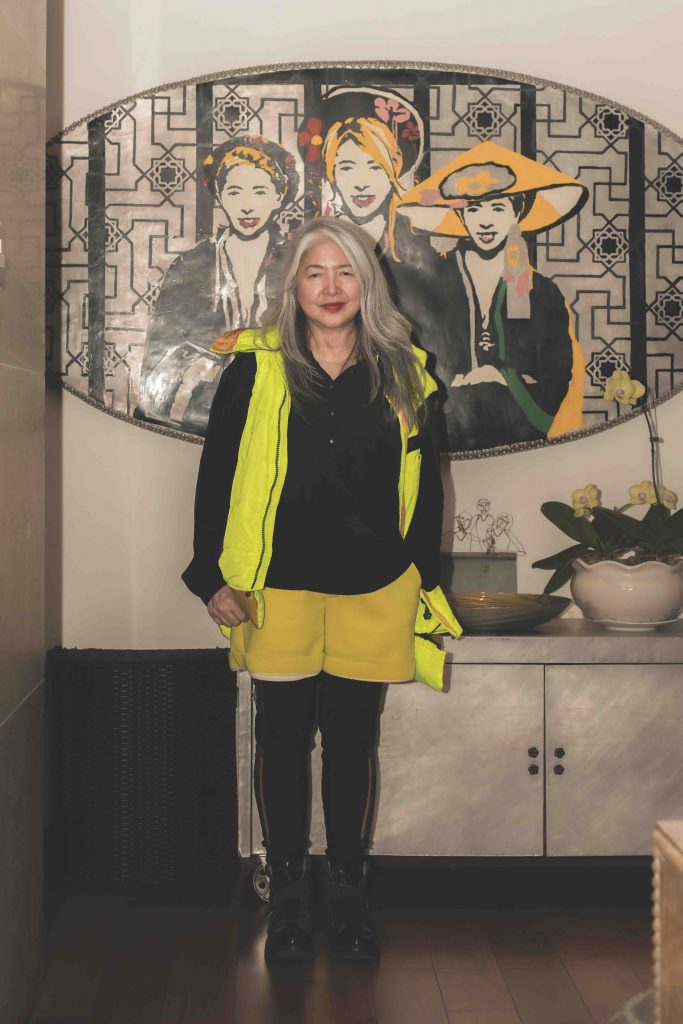 A woman in yellow stands before a colorful portrait and white patterned wall