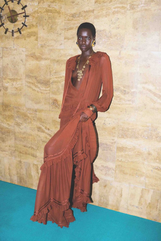 A model poses in a flowing rust colored Saint Laurent dress and gold jewelry