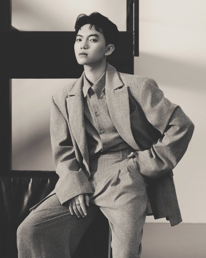 A young man sits wearing a textured gray suit and looks off to the side
