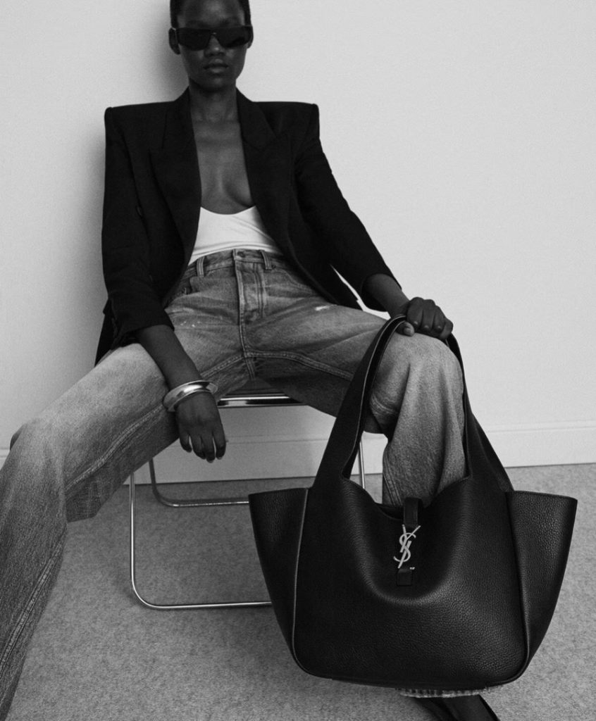 A model wearing sunglasses a blazer and jeans holds a large black tote bag