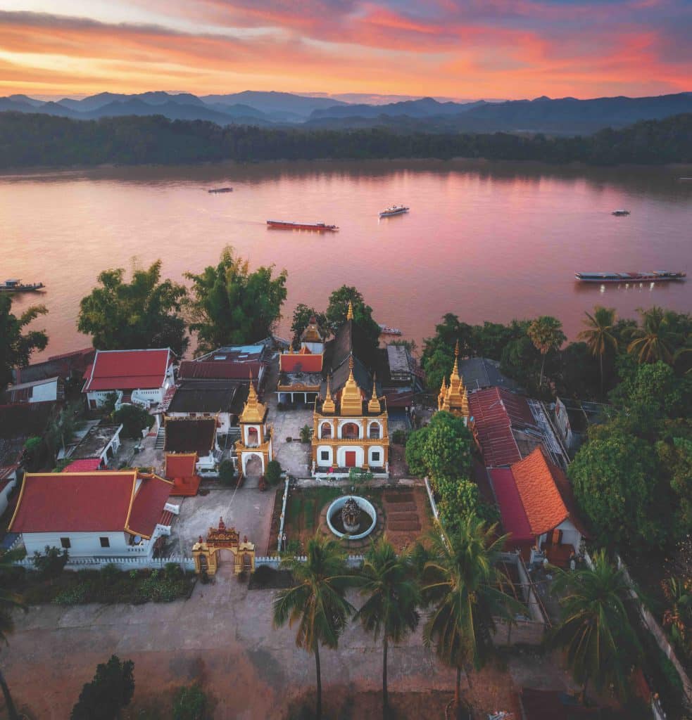 Golden temple roofs shine near a wide river at sunset with distant mountains