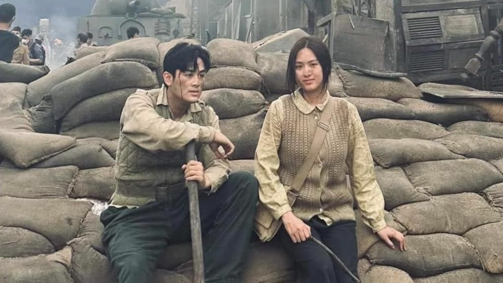 Cặp đôi diễn viên mặc trang phục thời chiến ngồi giữa bối cảnh chiến tranh với bao cát